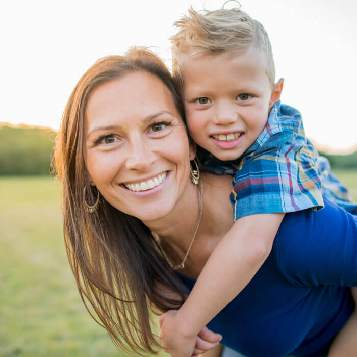 mother smiling outdoors with son hanging on her back