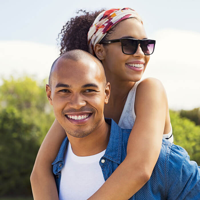 man smiling outdoors with woman in sunglasses hanging on his back